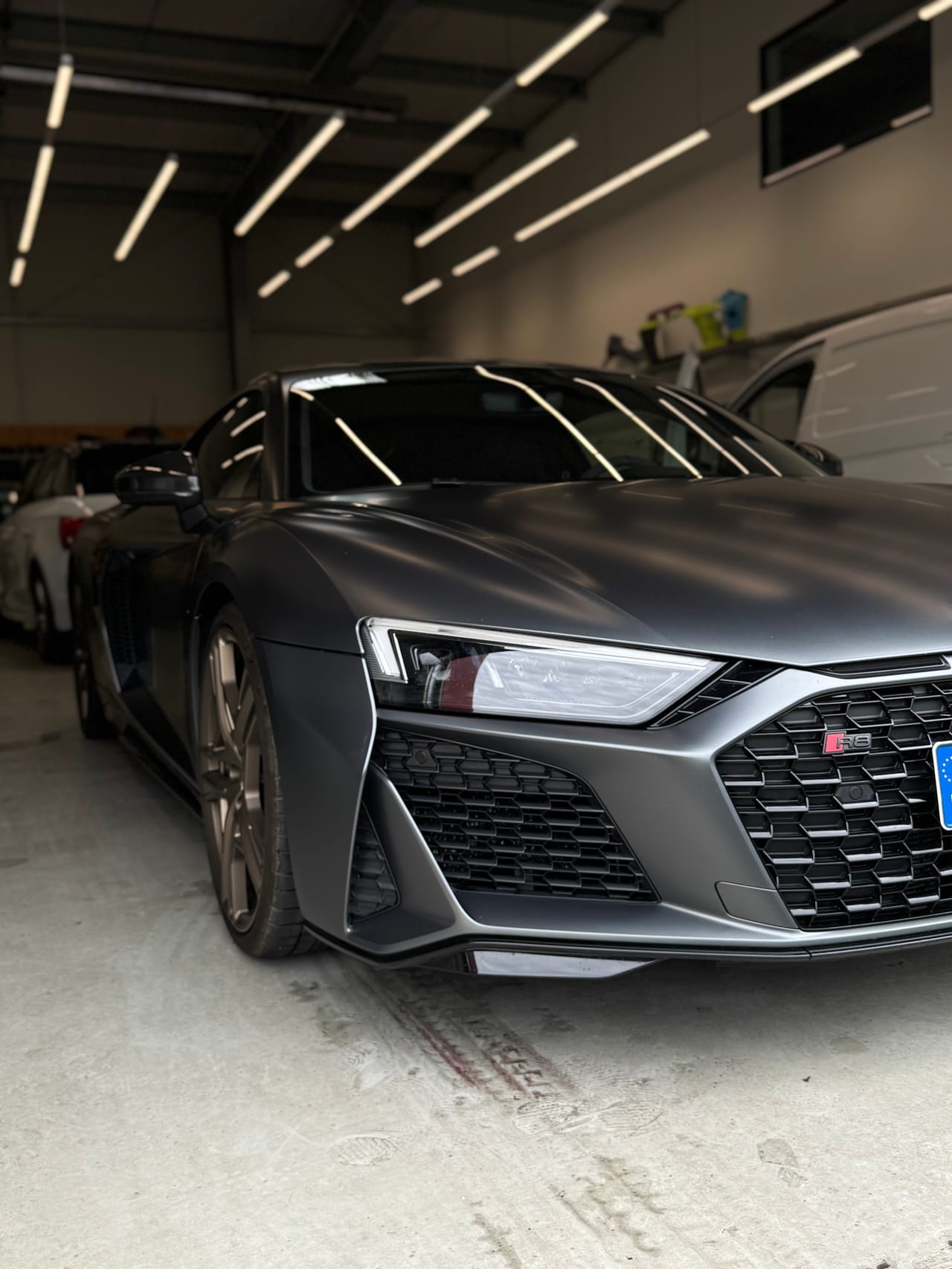Mattgrauer Audi R8 in der Aufbereitungshalle, von schräg vorne aufgenommen unter LED-Lichtleisten.