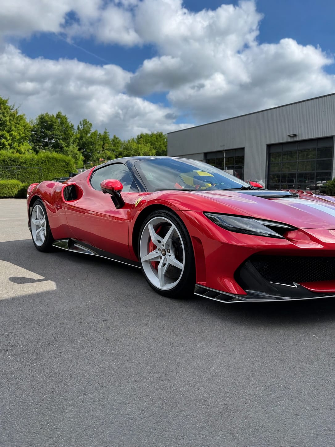Roter Ferrari 296 von schräg vorne im Freien vor einer Halle unter blauem Himmel, frisch aufbereitet und glänzend.