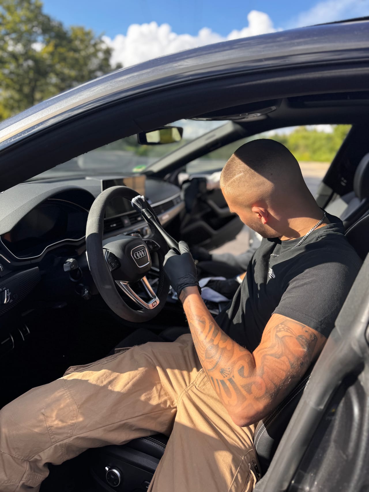 Jonas Braun reinigt mit Handschuhen das Lenkrad eines Audi RS im Freien unter blauem Himmel.