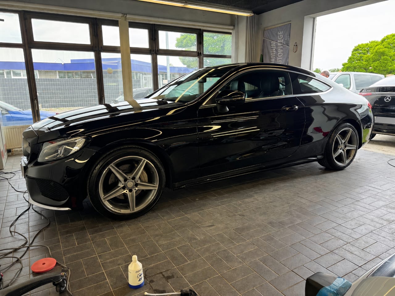 Seitenansicht eines schwarzen Mercedes C-Coupé in der Halle, davor Poliermaschine und Politur als Hinweis auf die Lackaufbereitung.