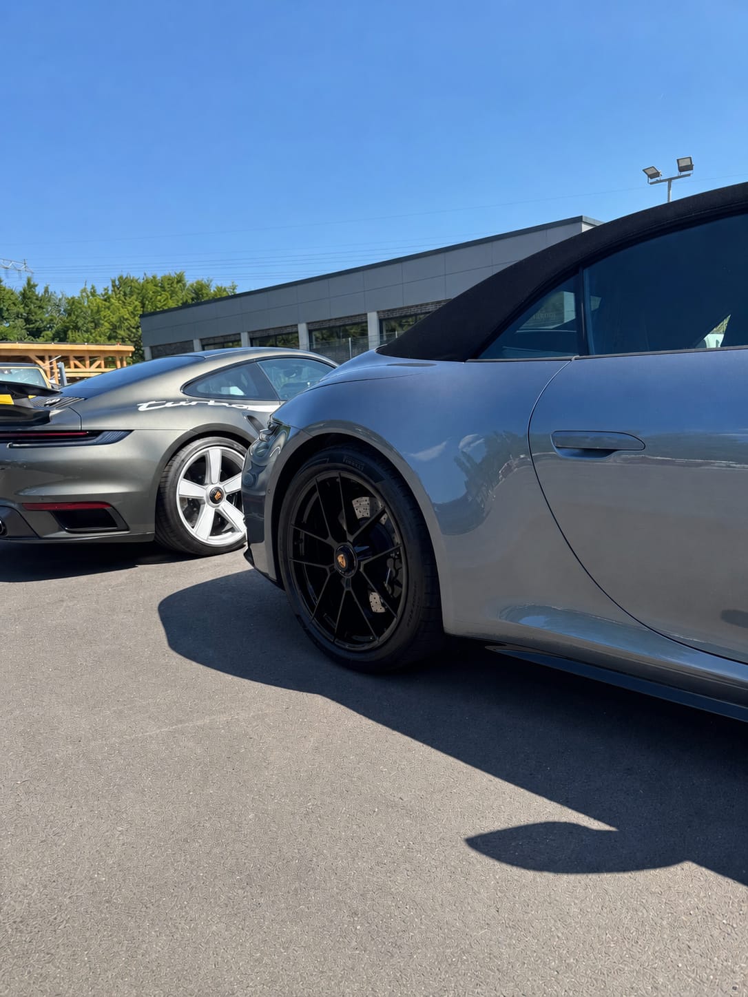 Zwei frisch aufbereitete Porsche 911 im Freien unter blauem Himmel: graues Turbo Coupé und graues Cabriolet mit schwarzem Verdeck.